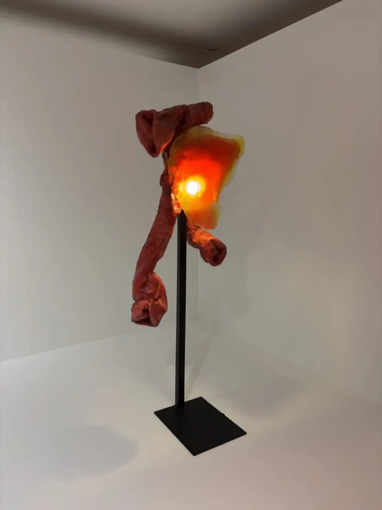 An orange mixed media sculpture resembling tendrils made with foam, with a light nestled in the center positioned on a display stand.