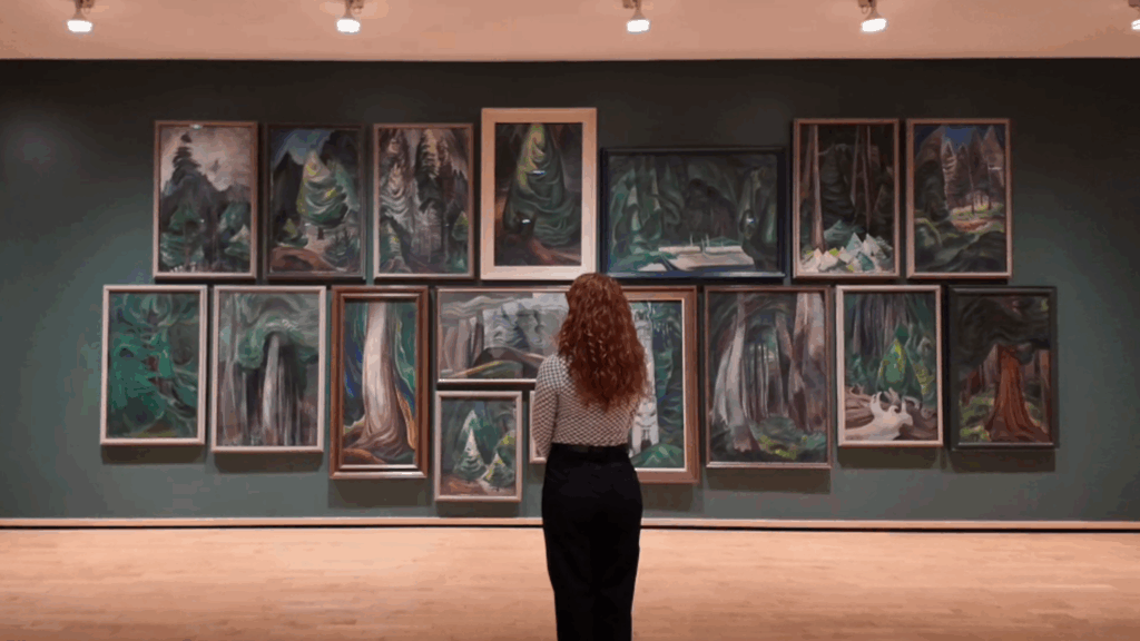 A woman standing in front of a selection of Emily Carr paintings hung on a wall in a salon style.
