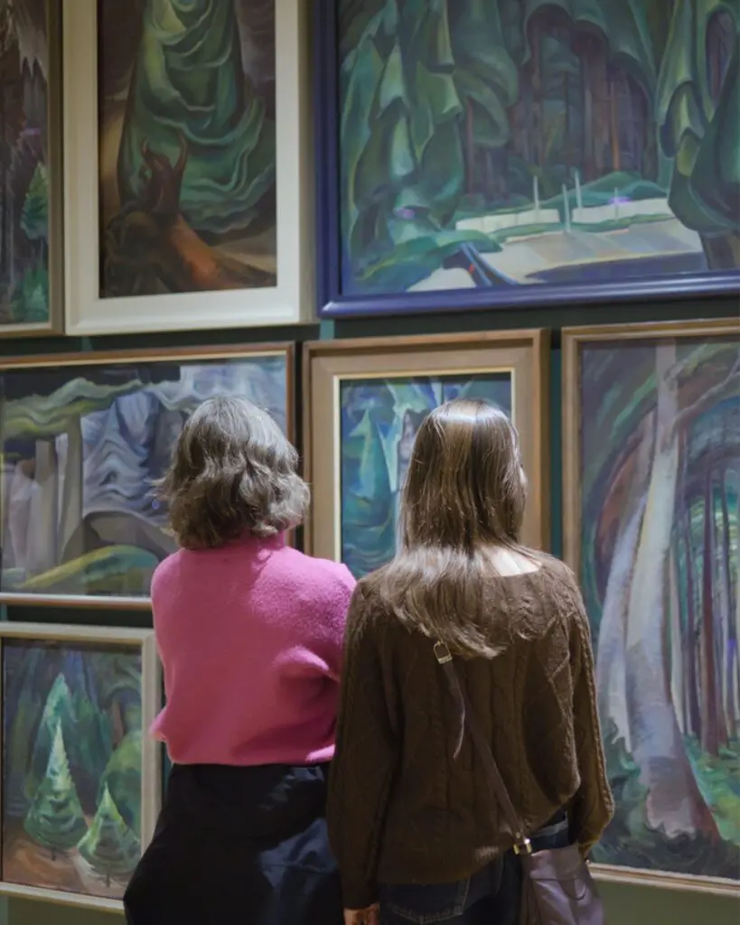 Two visitors looking at paintings by Emily Carr hanging in a salon style.