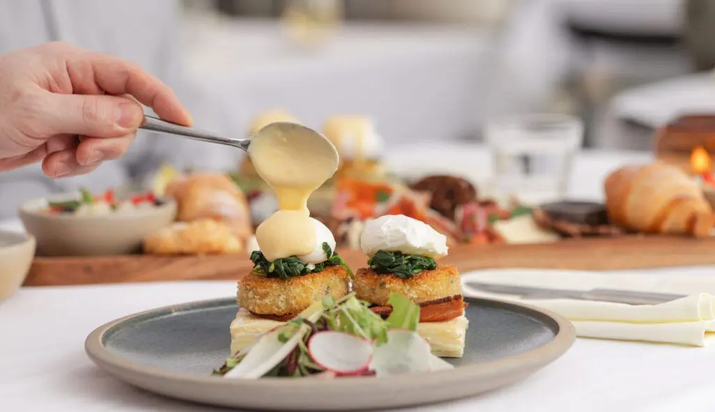 A hand drizzles hollandaise sauce over a plate of eggs benedict.