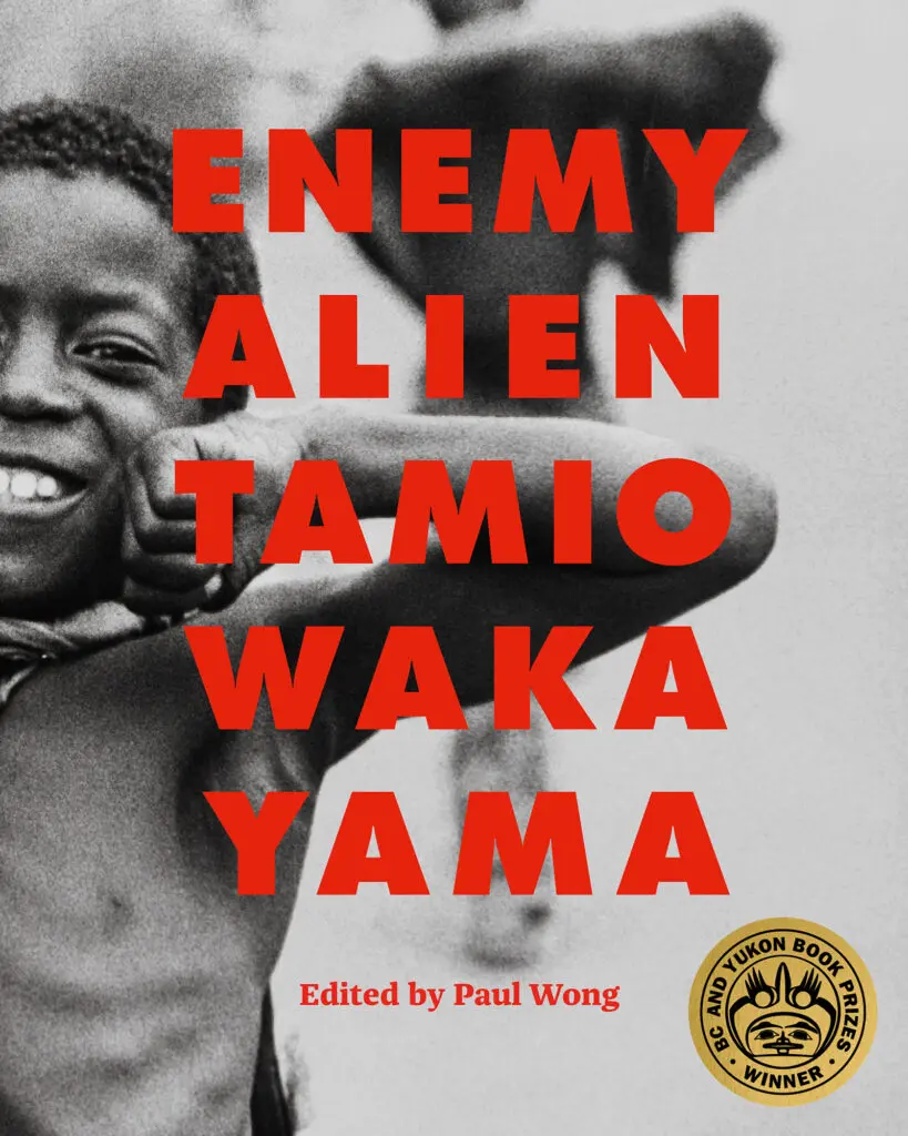 Cover of an exhibition catalogue with red, capital letters that read "Enemy Alien Tamio Wakayama." Below the title, in a smaller font, it reads: "Edited by Paul Wong." The red letters appear over a black-and-white photograph of a young, black boy with no shirt on, who smiles and beams out at the camera, while seemingly flexing his muscles. There is also a golden seal added to mark the book as winner of the winner of the Roderick Haig-Brown Regional Prize at the 2026 BC and Yukon Book Awards.