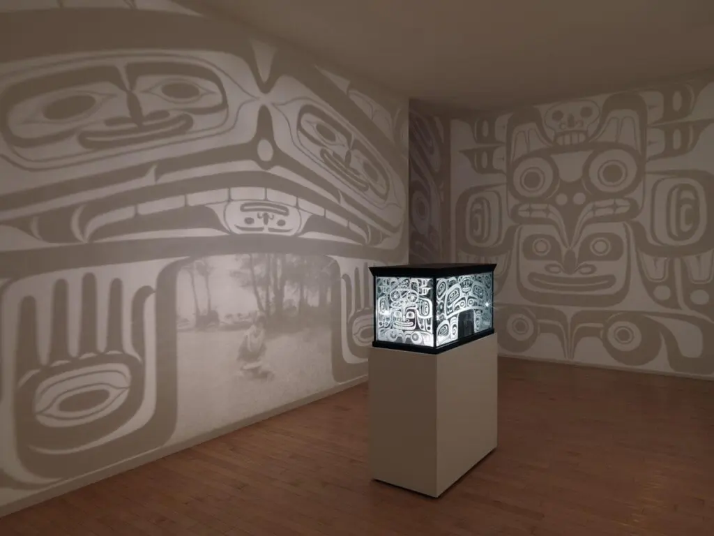 Installation by Marianne Nicolson with an object on a plinth in the centre of a room. The object looks like a bentwood box, and it shines light out on to the walls around it.
