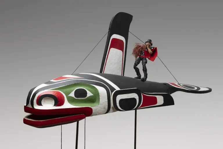 Photograph of an Indigenous installation artwork by Beau Dick of a large wooden whale with a small doll-like figurine of the artist on the whale's back.