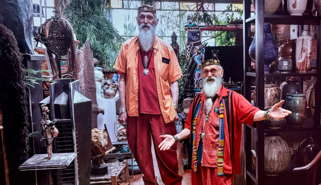 John David Lawrence, a white man with a long white beard, stands in from a mural photograph of himself in the same orange outfit. In the mural, he is standing in his home surrounded by ceramics on shelves.