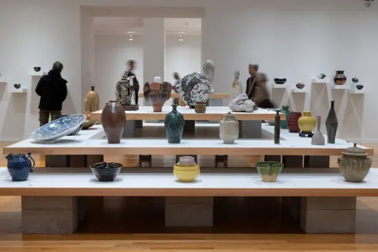 An installation view of Written In Clay. It looks at 3 shelves from the side. The shelves have multiple colourful vases and ceramics on them.