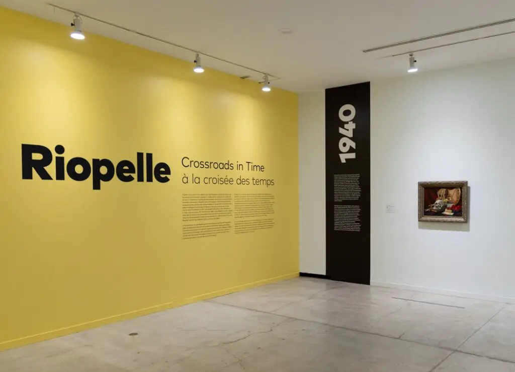Installation view of the exhibition "Riopelle: Crossroads in Time," with the title wall on a bright, yellow background, and a black vertical banner on the wall with the date 1940. On the wall adjacent to the title wall, there is a painting of a skull still life.