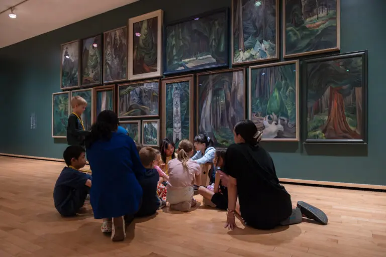 A group of children and adults sit in the Gallery in front of a wall of Emily Carr paintings of trees on a tour.
