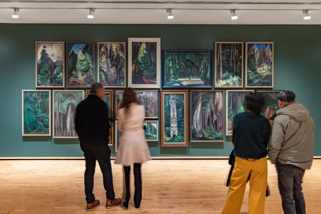 Two pairs of men and women stand in front of a wall of Emily Carr forest paintings