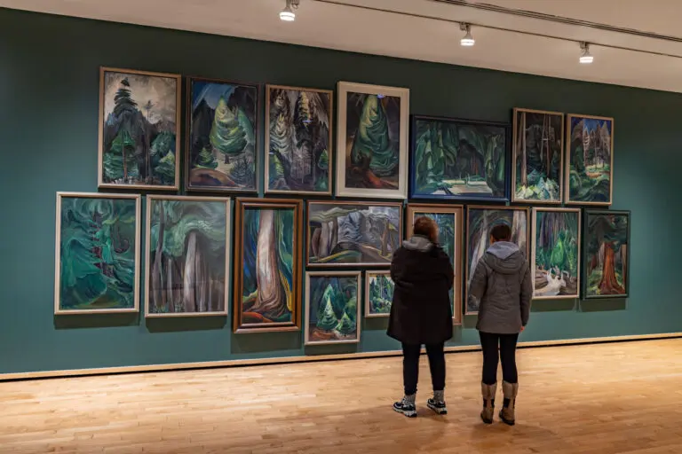Two women stand in front of a wall of Emily Carr forest paintings.