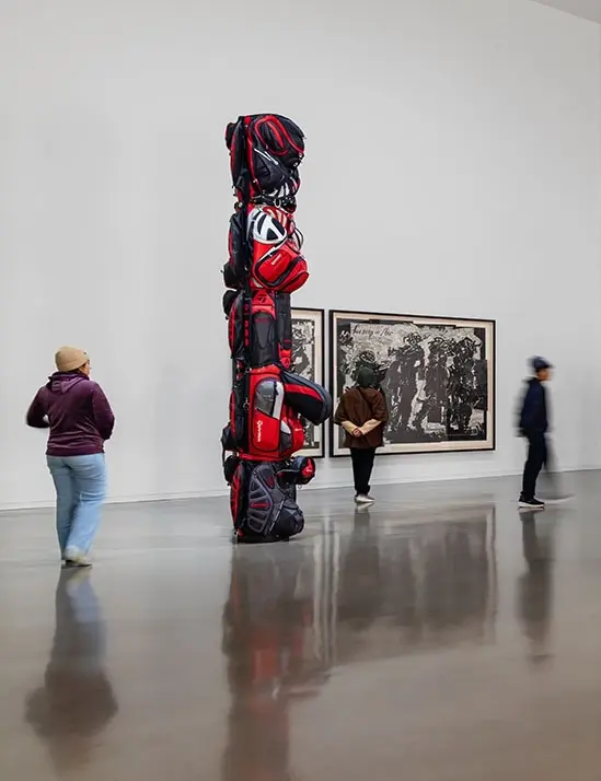 Installation view of the exhibition Postcards from the Heart: Selections from the Brigitte and Henning Freybe Collection. A sculpture that looks like a totem pole is visible in the foreground. It is made out of red golf bags. A woman stops to look at it, and two other visitors are visible in the background.