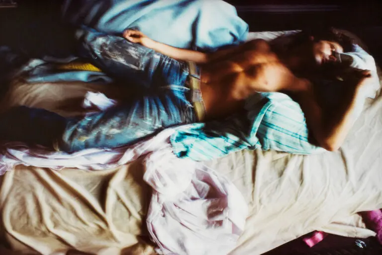 A slightly out-of-focus photograph of a shirtless man in denim blue jeans lying on a settee chair covered in wrinkled ivory and pastel blue sheets.