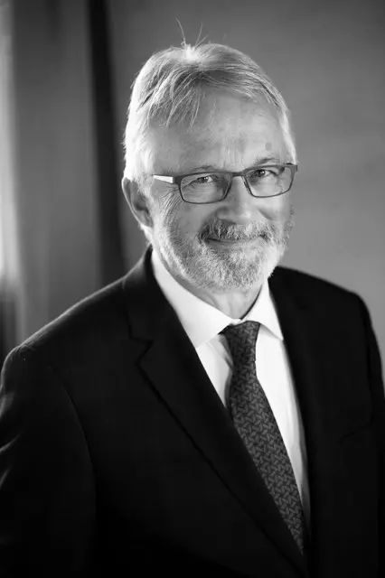 Black and white photograph of a man from the torso up, wearing a tie and a black suit jacket. He has white hair, a beard and wears glasses.