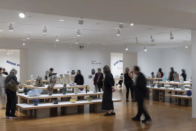 An installation view of Written In Clay. People walk around the room and discover platforms of shelves with many colourful vases and ceramics on them.