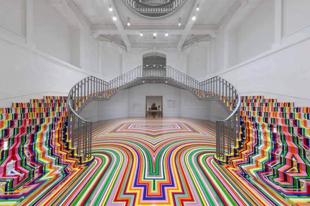 An installation photograph of Jim Lambie's artworks Zobop (Colour-Chrome) and Zobop (Colour-Chrome) Stairs covering the stairs and floor of the Vancouver Art Gallery's neo-classical Rotunda. The floor and stairs are covered in brightly coloured strips of vinyl in purple, pink, green, red, orange and yellow.