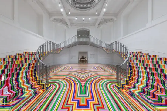 An installation photograph of Jim Lambie's artworks Zobop (Colour-Chrome) and Zobop (Colour-Chrome) Stairs covering the stairs and floor of the Vancouver Art Gallery's neo-classical Rotunda. The floor and stairs are covered in brightly coloured strips of vinyl in purple, pink, green, red, orange and yellow.