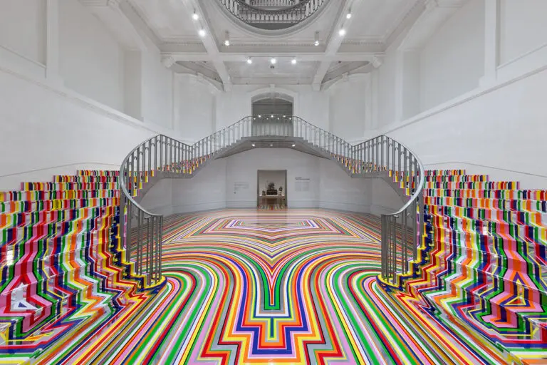 An installation photograph of Jim Lambie's artworks Zobop (Colour-Chrome) and Zobop (Colour-Chrome) Stairs covering the stairs and floor of the Vancouver Art Gallery's neo-classical Rotunda. The floor and stairs are covered in brightly coloured strips of vinyl in purple, pink, green, red, orange and yellow.