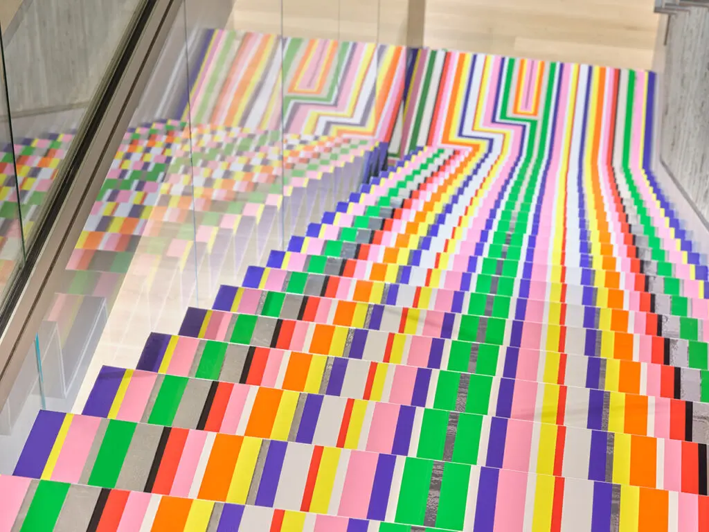 A detailed photograph of Jim Lambie's artwork on stairs. The shot is looking down a flight of stars that are covered in brightly coloured strips of vinyl in purple, pink, green, red, orange and yellow.
