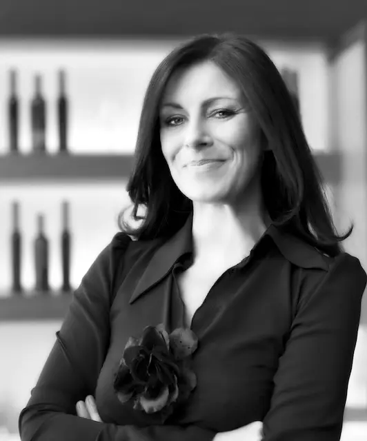 Black and white photograph of a woman with brown hair, smiling and standing with her arms crossed in a powerful pose, seen from the waist up. She is wearing a black suit jacket with a black flower on the front.