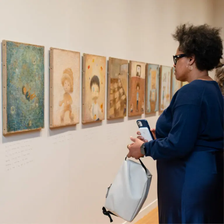 A Black woman wearing a blue dress and glasses stands on the right of the image and looks at paintings by Otani Workshop on the wall of the gallery.