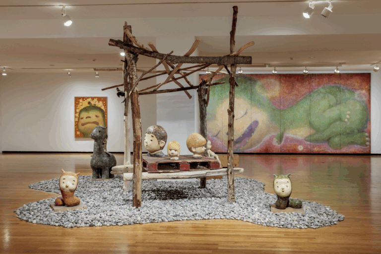 Installation view of the exhibition Otani Workshop Monsters in My Head, with rocks and a structure made of branches with two worm sculptures on either side and 2 boy head sculptures on the structure in the foreground. In the back, there are two paintings of Otani's green dinosaur-like character Tanilla.