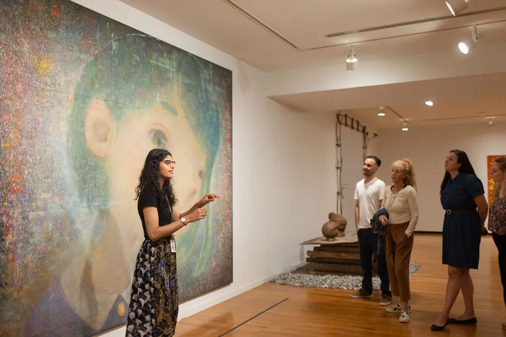 A young woman stands in front of a large painting of a girl on a wall. The woman is gesturing and speaking to a group of men and women onlookers.