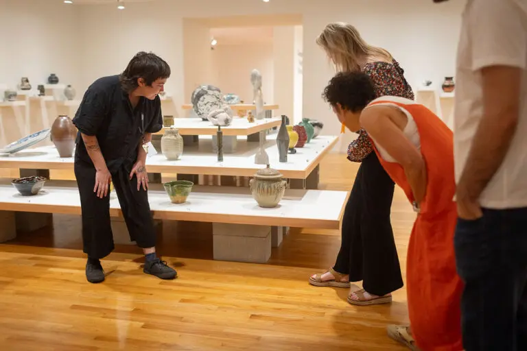 A group of men and women visit the Gallery and enjoy a guided tour. They are standing looking at shelves of ceramics as a tour guide speaks to them about the works.