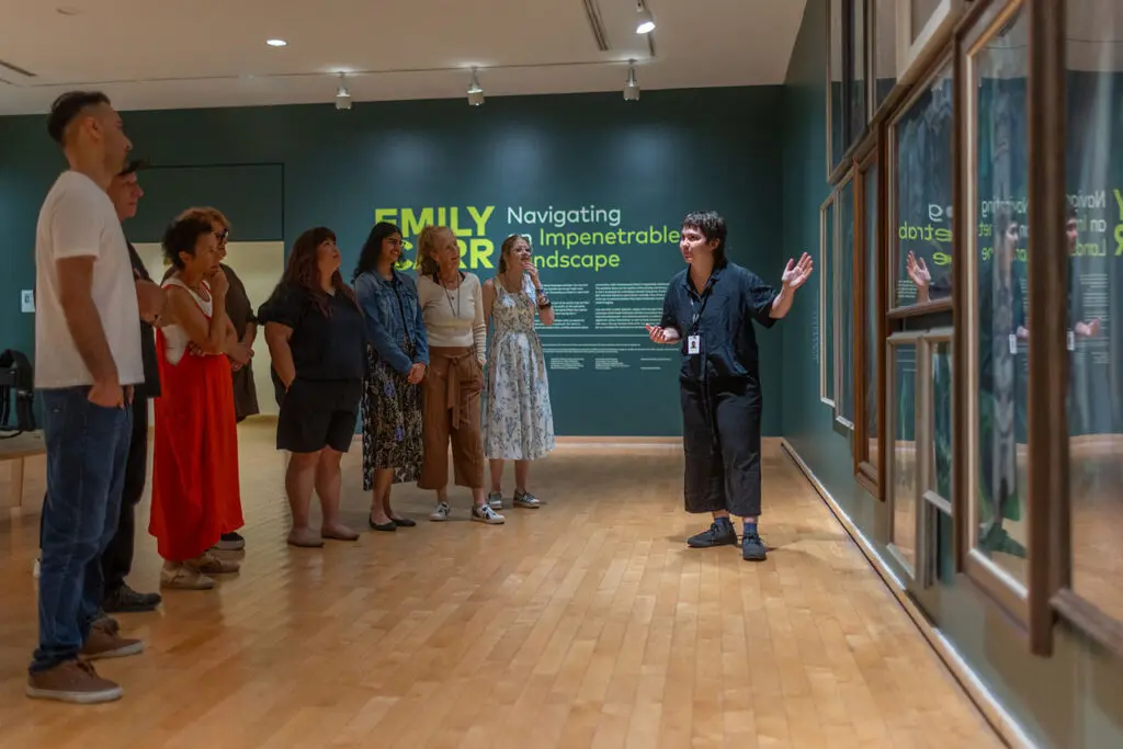 A group of men and women visit the Gallery and enjoy a guided tour. In this photograph, we can see them standing from the side, looking at a dark green wall with Emily Carr paintings of the forest on it. A young woman tour guide stands in front of the paintings gesturing and looks at the group.