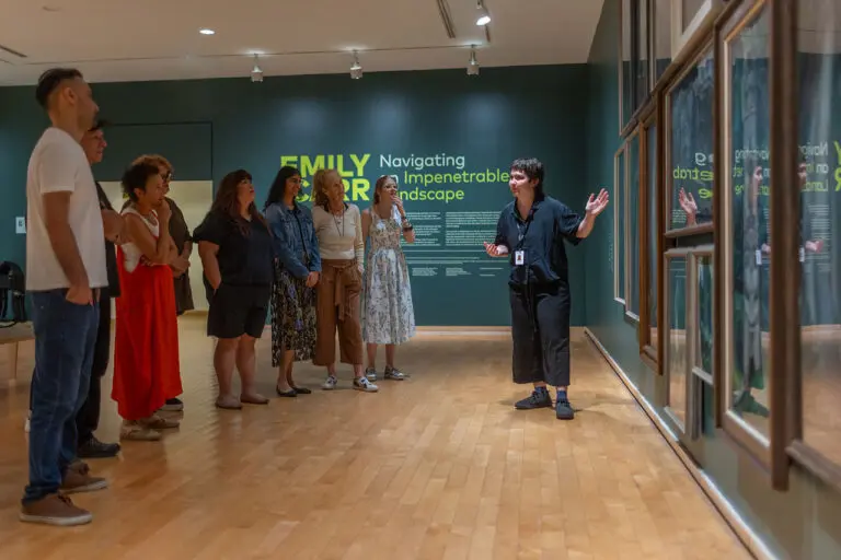 A group of men and women visit the Gallery and enjoy a guided tour. In this photograph, we can see them standing from the side, looking at a dark green wall with Emily Carr paintings of the forest on it. A young woman tour guide stands in front of the paintings gesturing and looks at the group.