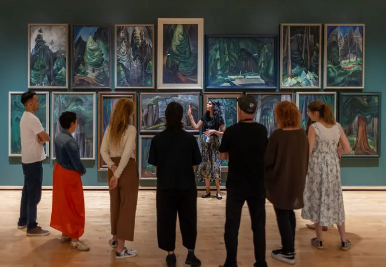 A group of men and women visit the Gallery and enjoy a guided tour. They are standing with their backs to us, looking at a dark green wall with Emily Carr paintings of the forest on it. A young woman tour guide stands in front of the paintings gesturing and looks back at the group.