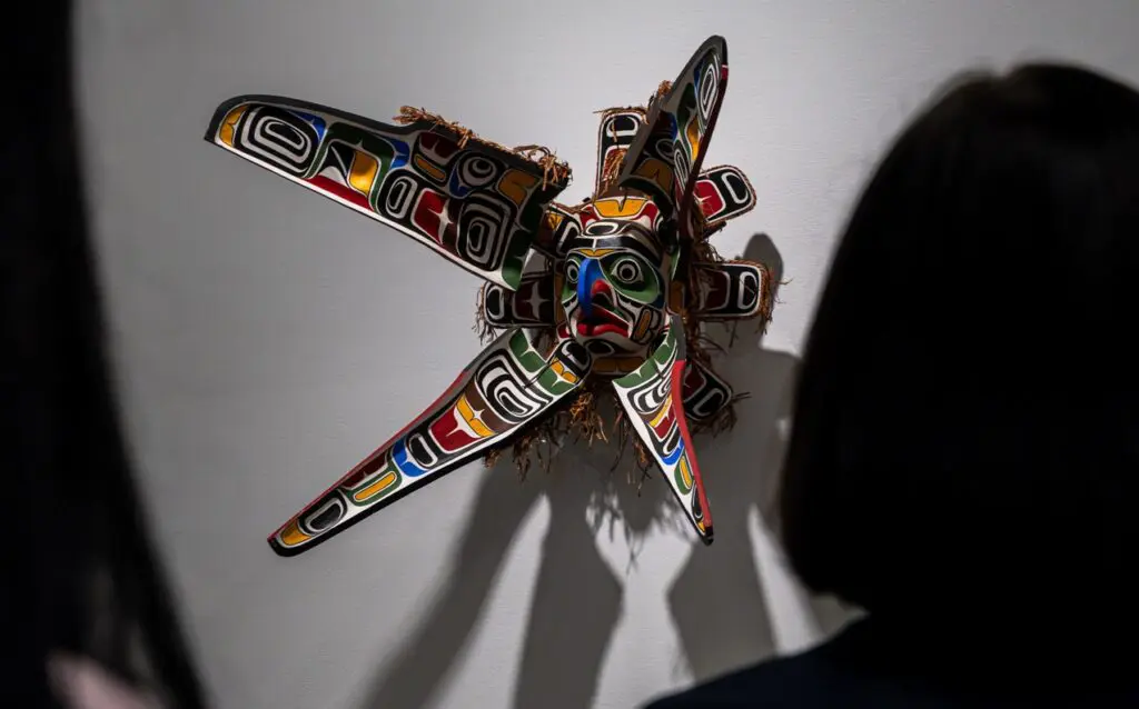 Two silhouettes of the back of women's heads appear in the foreground of the image, and they are looking at a Indigenous transformation mask by Beau Dick.