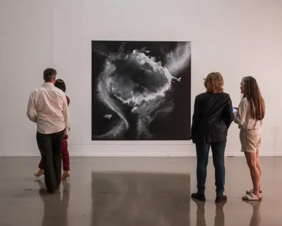 A photograph of 4 people (two women standing together and a man and a woman) from behind looking at Tacita Dean's pastel drawing of clouds in shades of black and white and grey.