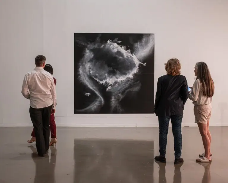 A photograph of 4 people (two women standing together and a man and a woman) from behind looking at Tacita Dean's pastel drawing of clouds in shades of black and white and grey.