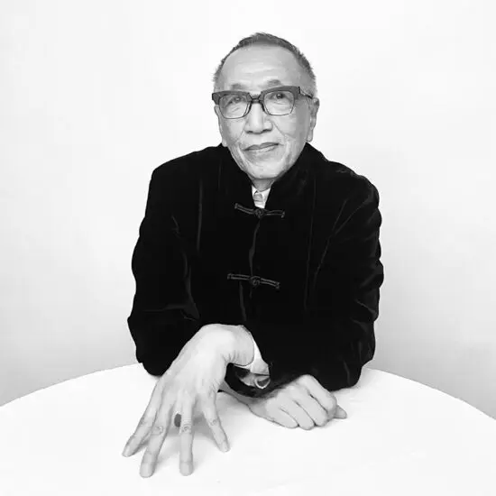 Headshot of artist and curator Paul Wong. In the black-and-white photograph, he is seen sitting at a round, white table. He is wearing black-rimmed glasses and a black jacket. His arms are bent, leaning on the table. One hand is raised, with fingertips touching the table.