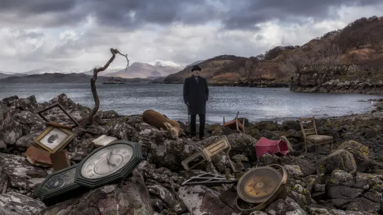 A film still featuring a man standing in a realistic yet surreal landscape. There are rocks and clocks in the foreground, and then a body of water and a mountain in the background behind the man. The landscape is pretty barren.