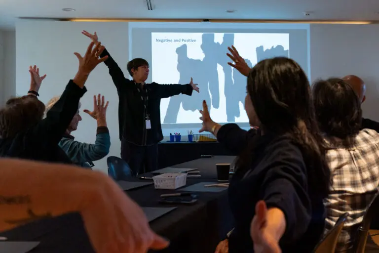 A photograph of a group of seniors, led by instructor Kathryn Ricketts standing in the centre of the image in front of a Powerpoint presentation. The seniors are sitting at tables with their arms outstretched as part of a movement workshop.