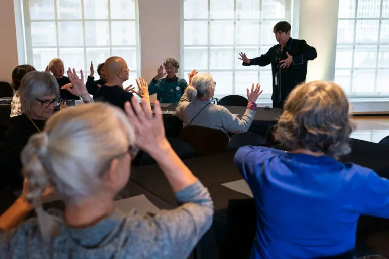 A photograph of a room with windows and tables and chairs with seniors looking at a presenter standing at the front of the room. Every one has their hands up and participates in a movement workshop.