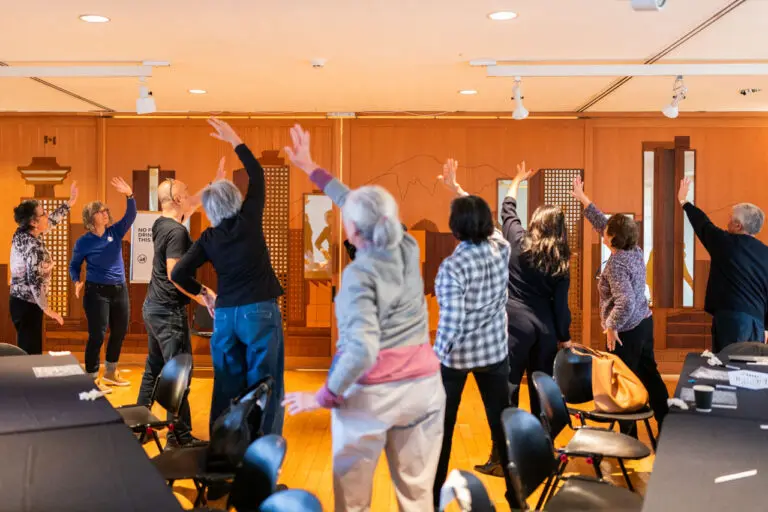 A photograph of a group of seniors from behind stretching their arms up during a movement workshop.