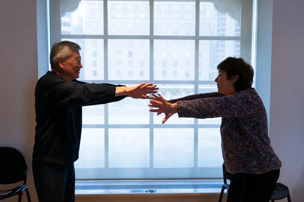 A photograph of two seniors from the side, one man on the left and one woman on the right. They are standing in front of a window grid with their arms stretched out towards each other, hands almost touching, as part of a movement workshop.