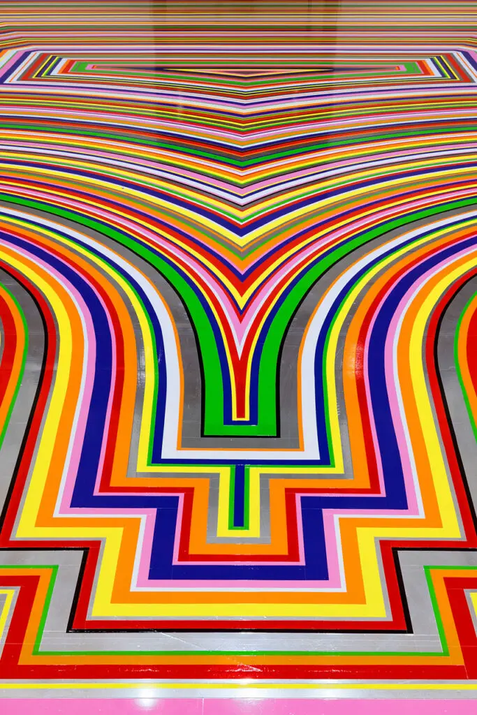 A detail photograph of Jim Lambie's artwork on the Gallery Rotunda's floor. The shot is looking at a detail of the floor covered in brightly coloured strips of vinyl in purple, pink, green, red, orange and yellow.