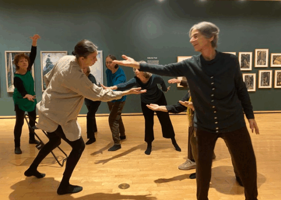 A photograph of a group of senior women dancing in an exhibition of forest paintings and drawings by Emily Carr.