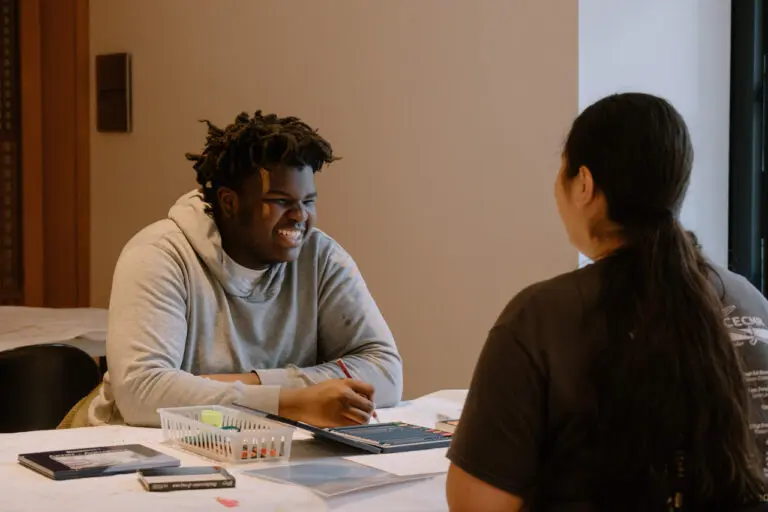 A man and a woman sit at a table making. We see the top of the table, the woman's back, and the man's face, smiling, as they create art together.