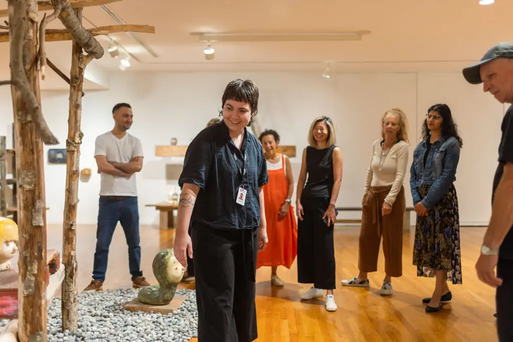 A group enjoys a tour of the exhibition Otani Workshop: Monsters In My Head, exhibition at the Vancouver Art Gallery, led by a woman in black with short brown hair, smiling.