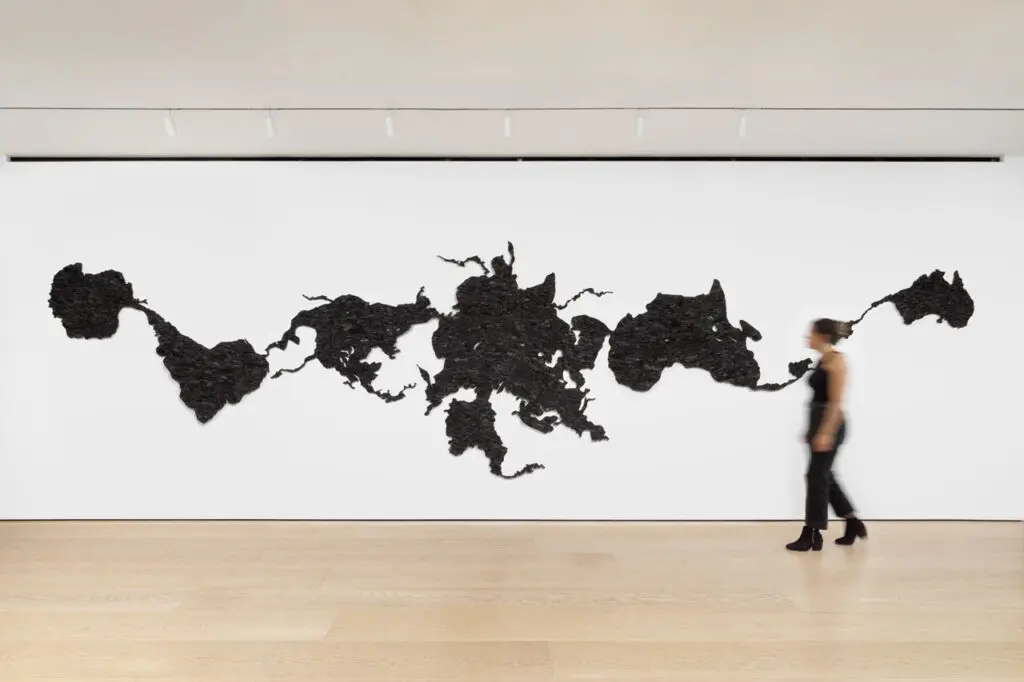 Installation view of Teresita Fernández's work Island Universe 2, made entirely of charcoal. The installation looks like a distorted map of the world, spread across one white wall. A blurred person walks in front.