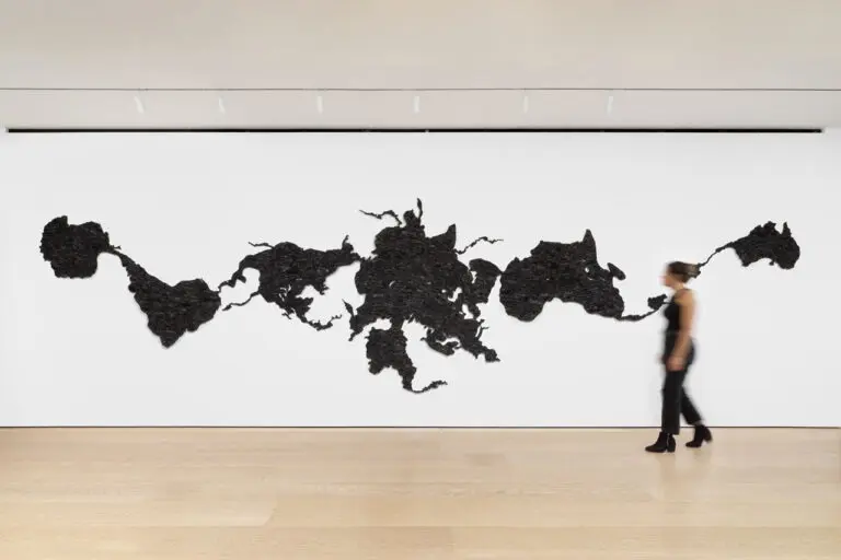 Installation view of Teresita Fernández's work Island Universe 2, made entirely of charcoal. The installation looks like a distorted map of the world, spread across one white wall. A blurred person walks in front.