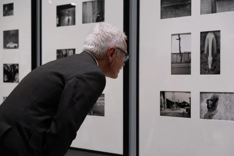A photograph of a white-haired man from the torso up, leaning in to take a closer look at a grouping of black-and-white photographs on the wall.