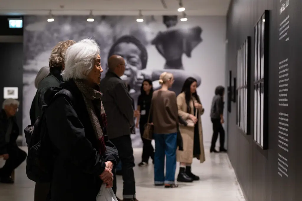 A photograph featuring an older Japanese woman from the side, looking at framed black-and-white photographs on a wall in front of her. People are mingling and looking at art around her in the background.