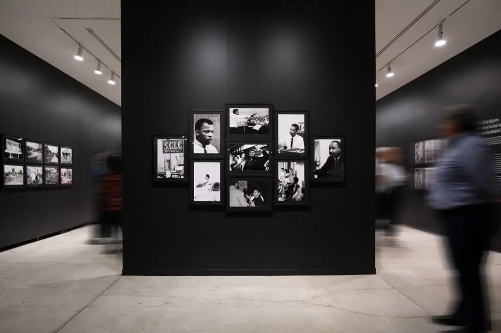 A photograph of a wall in the exhibition Enemy Alien: Tamio Wakayama. The wall colour is black. We see some moving people walking by. On the wall, there is a grouping of black-and-white photographs of important Civil Rights Movement figures.
