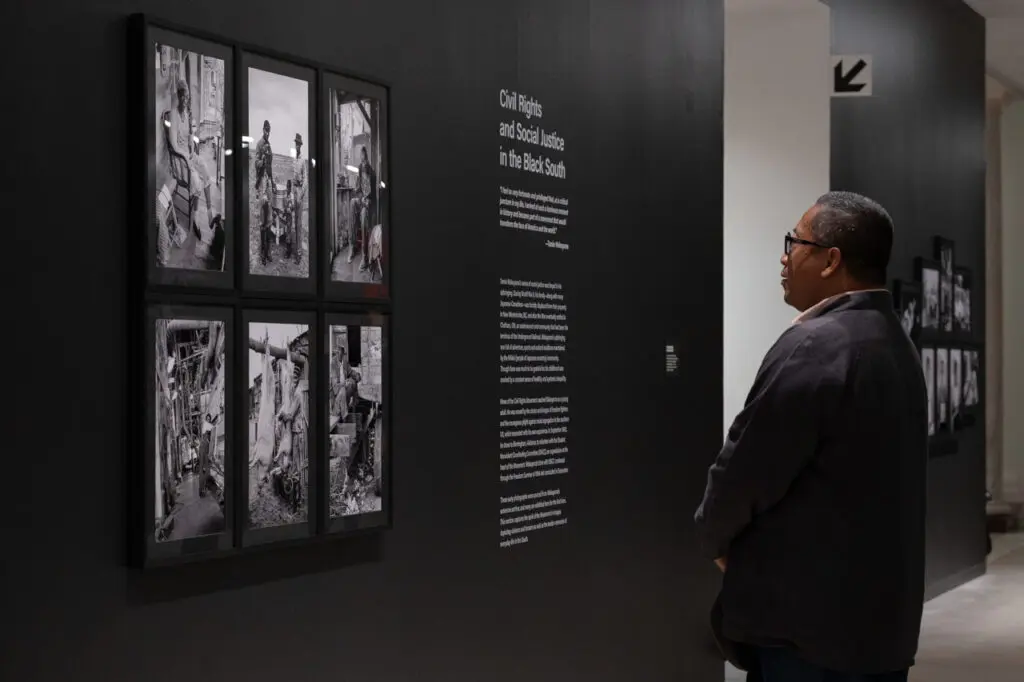 A photograph of a man standing and looking at a black wall with a series of 6 framed black-and-white photographs on it