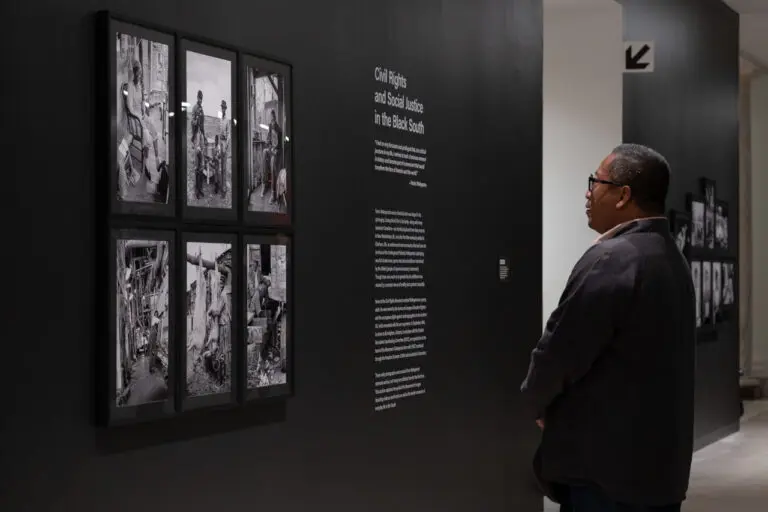 A photograph of a man standing and looking at a black wall with a series of 6 framed black-and-white photographs on it