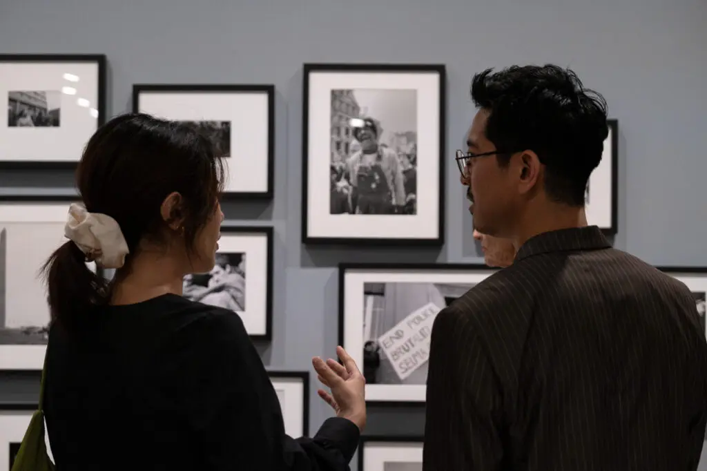 A photograph of two young Asian adults from behind, looking at each other and discussing the multiple black-and-white photographs on view behind them